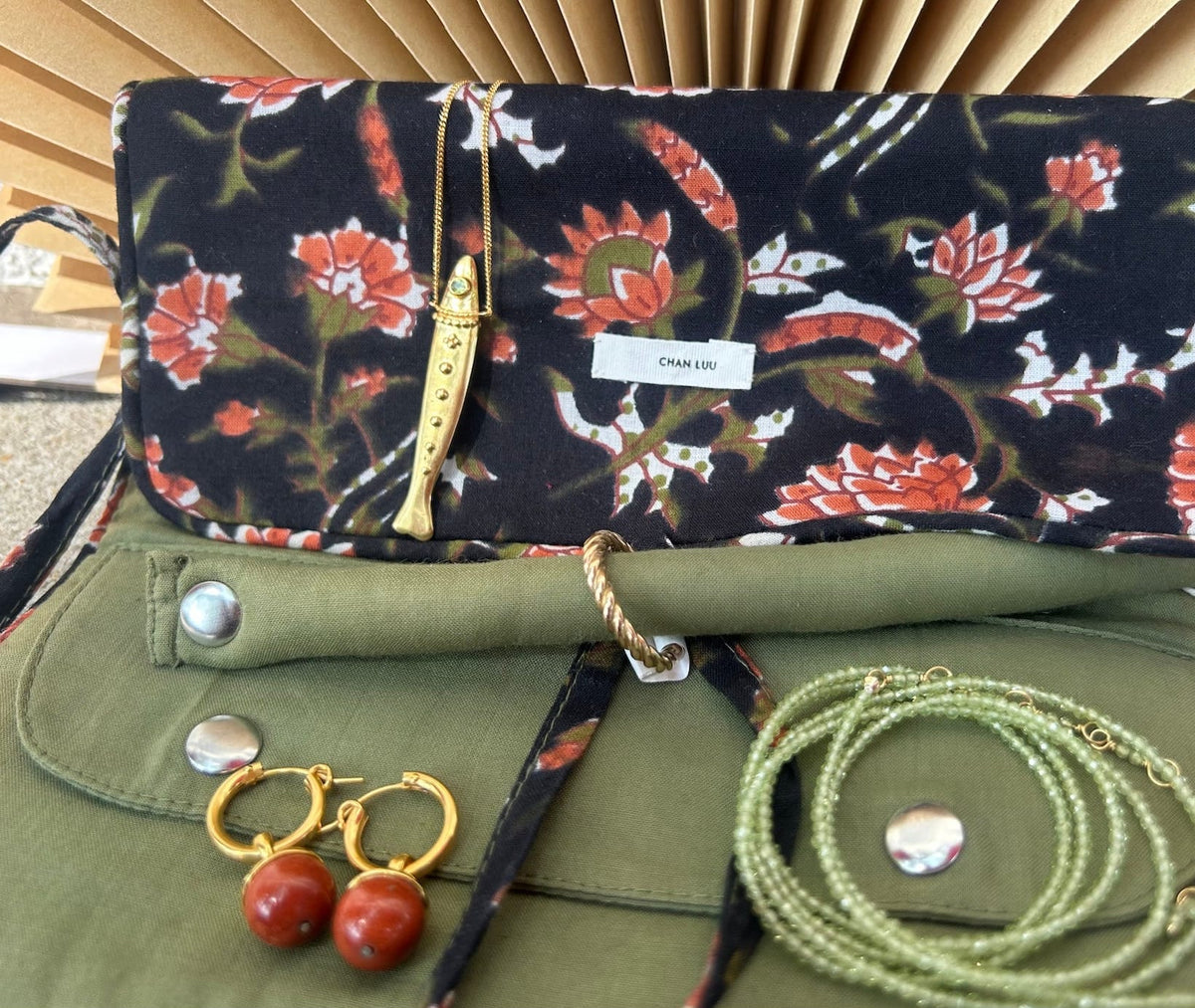 A floral fabric clutch bag with a black base and red, orange, and white flower print, styled with jewelry accessories on an olive green surface in front of a beige pleated background.