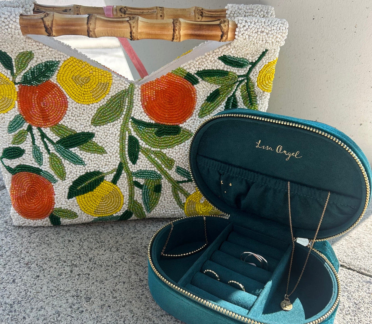 A teal velvet jewelry organizer case with gold trim, shown open with rings and a necklace inside, next to a white beaded handbag with orange and yellow fruit motifs and a bamboo handle.