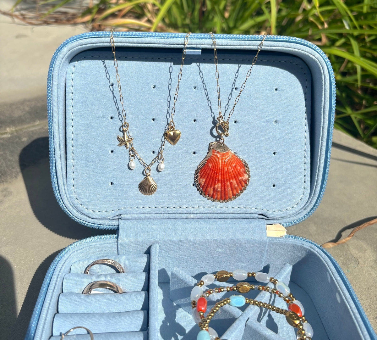 An open light blue jewelry organizer case displayed outdoors on a stone surface with green grass in the background. The case contains several necklaces and bracelets, including a coral shell pendant necklace, silver hoop rings, and beaded bracelets.